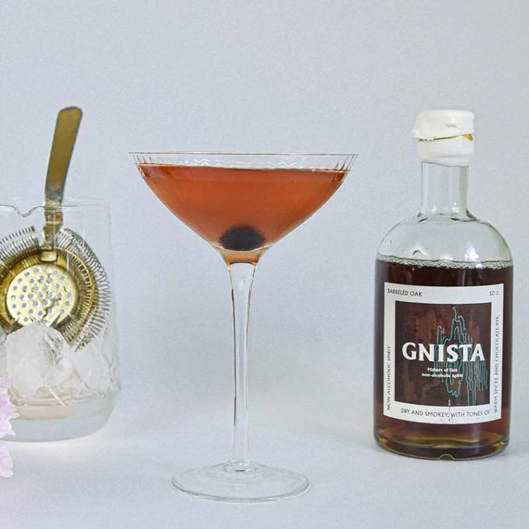 En elegant alkoholfri Gnista Manhattan serverad i ett martiniglas med ett maraschinokörsbär i botten, stående bredvid en flaska Gnista Barreled Oak och ett rörglas med is och barsil.