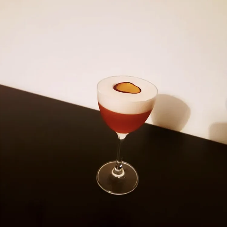 n röd-orange alkoholfri Grilled Plum Paper Plane-cocktail med ett tjockt lager vitt skum, garnerad med en vacker karamelliserad plommonskiva som flyter på toppen i ett elegant glas på fot mot en mörk bänkskiva.
