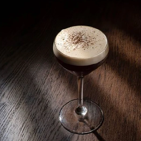 En stämningsfull alkoholfri kaffecocktail i ett elegant coupeglas, med ett tjockt lager ljust skum toppat med krossade kaffebönor, placerad i dramatiskt ljus på ett mörkt träbord.