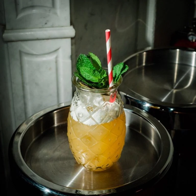 En gul alkoholfri Mai Tai-inspirerad mocktail serverad över is i ett ananasformat tikiglas, garnerad med en generös myntakvist och ett rödvitt papperssugrör, stående på en rund metallbricka.