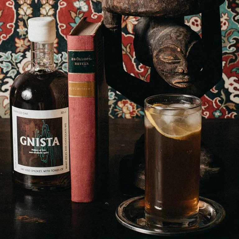 En hög alkoholfri Oak Tonic serverad med is och en citronskiva i ett highballglas på ett silverfat, stående bredvid en flaska Gnista Barreled Oak, en äldre bok och en mörk träskulptur mot en mönstrad blommig bakgrund.