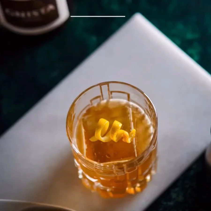 En alkoholfri Gold Rush mocktail serverad med en stor iskub och en snurrad citrontwist i ett mönstrat kristallglas, stående på en vit marmorskiva mot en mörkgrön bakgrund.