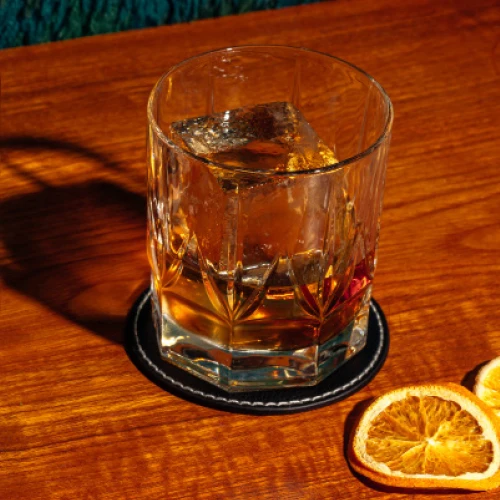 En stämningsfull alkoholfri Old Fashioned serverad med en stor klar iskub i ett kristallmönstrat whiskeyglas på ett mörkt underlägg, fotograferad i varmt ljus på en träskiva med torkade apelsinskivor intill.